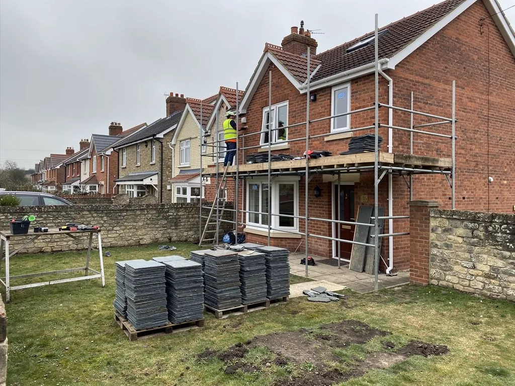 Pallets of new slates in a garden beside a scaffolded house