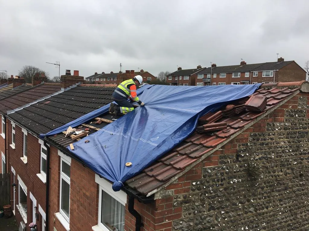 Roofer securing a blue tarpaulin over storm-damaged roof tiles